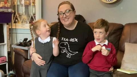 Debbie McCrudden sat on a sofa with her daughter and son both in school uniform 