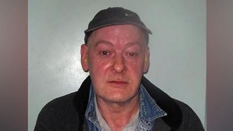 A custody-style photograph showing a Sweeney wearing a dark cap, denim shirt layered under a dark jacket and a light-coloured top, standing against a plain light background.