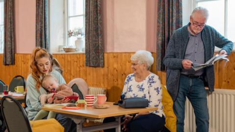 A woman and a baby are on a chair in the left of the image. At the right is a woman in a white flowery top siting down and stood next to her is a man reading a paper. On the table are mugs and biscuits. 