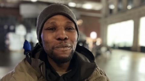Josh looks into the camera. He is wearing a black hat and a grey-green coat over the top of a black hoody. Behind him, out-of-focus, are shops in the centre of Leeds