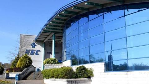 A large, curved glass-fronted building with a covered entrance to the left and the letters 'NSC' on the left-hand wall approaching the entrance. Stairs lead up to the entrance with shrubbery to the right and left.