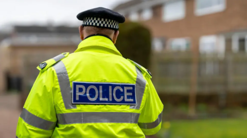 A photo of a police officer from behind. They are wearing a high-vis jacket which reads Police. 