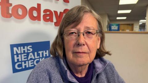A woman wearing glasses in a blue fleece in front of an NHS sign
