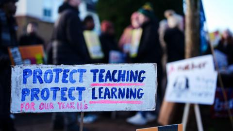 Signs belonging to members of the National Education Union on a picket line outside the Arthur Terry Learning Partnership (ATLP) school in Sutton Coldfield. The image shows a white rectangular sign made out of cardboard. The sign is painted with blue and pink lettering which reads: "Protect teaching, protect learning for our Year 11s."