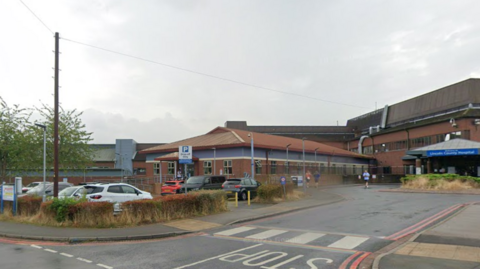 Exterior shot of the main entrance to Lincoln County Hospital. It is a red-bricked building with a blue sign over the main doors. There are other buildings in the background, as well as parked cars.