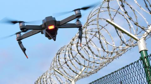 A black drone with an orange light overs over a barbed wire fence on a sunny day