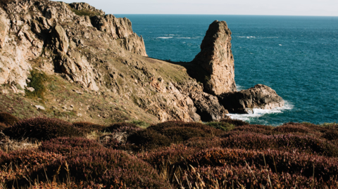 A picture of Le Pinacle in Jersey. It is a rock next to the ocean. the water is a dark blue and the sky is grey. There are brown bushes in the forground and a rocky hill in the background. 