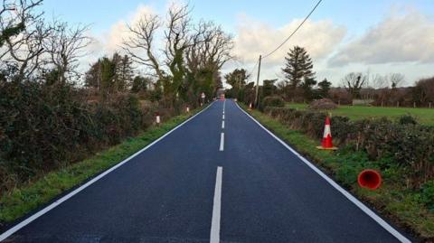 A newly resurfaced road stretching out into the distance. The carriageway is black with bright white lines painted on it. There are green hedgerows and fields on either side and traffic cones to the right.