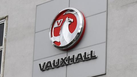 The red and silver logo of Vauxhall on the side of the grey factory building in Luton