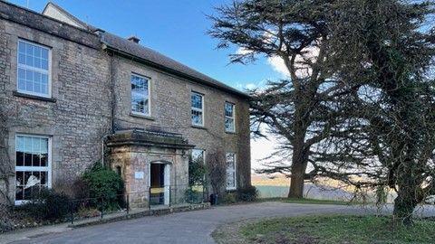 The Wilderness Centre, a country home in the Forest of Dean, with a large tree in front of it and a turning circle. Panoramic views across the Forest can be seen behind it on what is a sunny day.