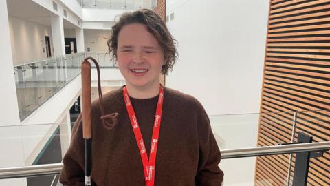 A girl with long brown hair wearing a brown jumper and a red University of Northampton lanyard stands in a stairwell holding a cane.