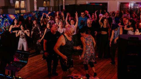 A dance hall filled with people of all different ages, some dressed in colourful outfits and even face paint, many with their arms up in the air.