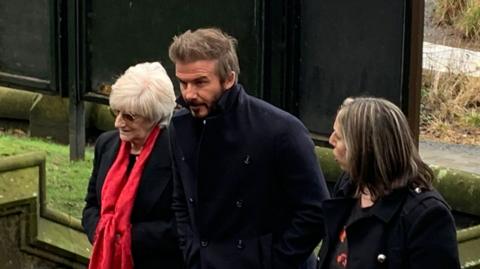 David Beckham arriving at Manchester Cathedral wearing a dark coat and flanked by two women