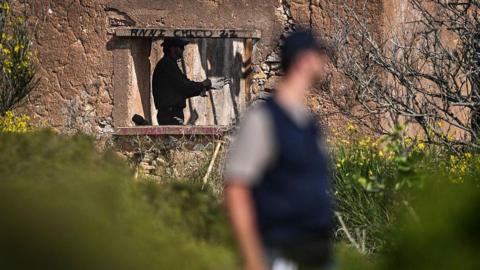 Derelict building with a man standing inside it, visible through the hole in the wall where a window would have been. On the upper frame is black graffiti. A blurred man is standing in the foreground to the centre-right of the picture. There is vegetation surrounding the building.