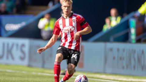 A blond footballer in a red and white striped jersey, black shorts and red socks dribbles the ball on a sunny day at a small stadium. His shorts have the number 14 on them.