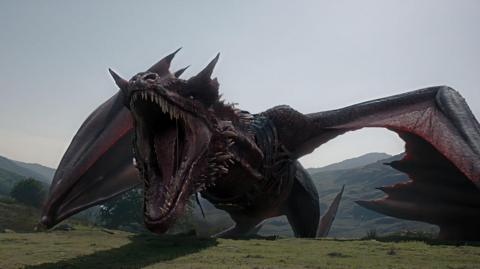 The head and neck of a large, red, computer-generated dragon rises over the top of the mountain; it is roaring, revealing sharp teeth with its wings outspread