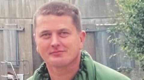 A smiling Paul Ainscough, with short cropped brown hair and wearing a green jacket, stands in front of a grey concrete wall and a green bush