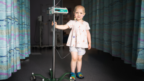 A photo of a little girl standing in the camera, she is wearing shorts and a top and holding onto a machine.