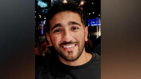 An image of a man with short dark hair, a moustache and beard smiling at the camera. He is wearing a black jumper and appears to be standing in a nightclub.