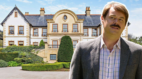 The entrance to a mansion. The property is a beige and white colour with perfectly trimmed bushes in front of the house. A circular driveway can be seen looping around the entrance to the property. And there is also a photoshopped image of Danny Dyer on the image.
