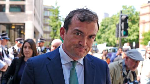 Patrick Spencer leaves a court, dressed in a blue suit and green tie. He is smiling at the cameras. In front of him is a police officer, dressed in uniform.
