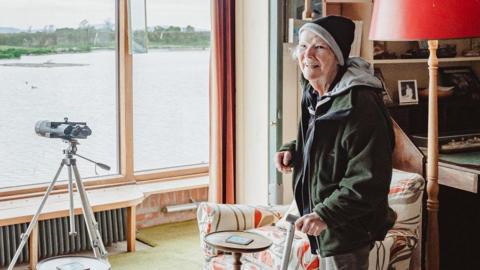 An elderly woman in a hat stands by a large window with a telescope in front of it