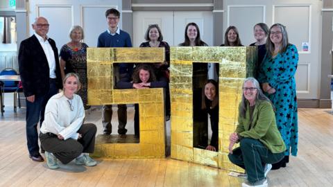 Ten woman and two men stood next to a giant gold 50 sign that comes up to about shoulder height. All are looking at the camera and smiling. They are inside a large hall with wooden floors and paneled walls.