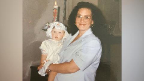 Rochelle Crane, holding her daughter Louise Crane. Rochelle is wearing a light blue shirt and has round spectacles. 