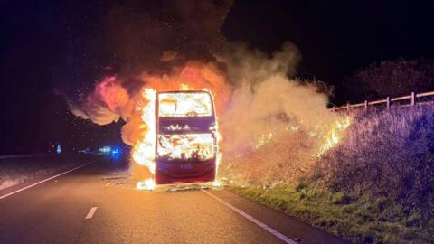 A bus fully engulfed in flames with smoke plumes coming from it. The sky is dark and it is on the side of the road. 
