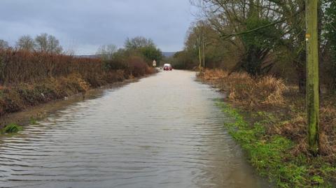 A long narrow stretch of flooded road lined with trees and hedgerows. In the distance, firefighters in orange waders are guiding a raft with a person on board away from a partly submerged white van.