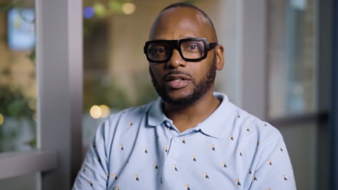 Christopher Joell-Deshields looks at the camera, his lips pursed as though he is about to speak. He is wearing a blue polo shirt covered in embroided puffins. The background is out of focus