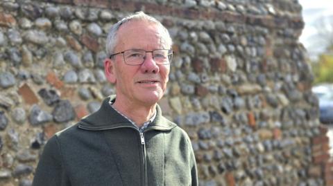 Brian Watkins is wearing a green cardigan over a blue and white checked shirt. 