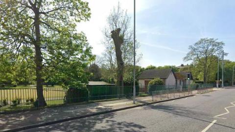 Google streetview of bowling green at Birkenhead Park behind the railings next to a road. It is surrounded by trees and a one-story building near a traffic light crossing.
