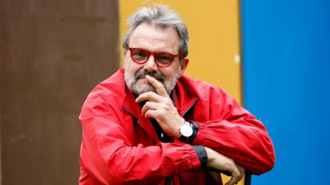 Master photographer Oliviero Toscani poses for photographers in a red jacket with a brown, yellow and blue backdrop. 