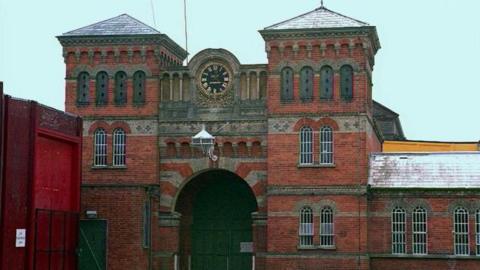 The front of a red brick building with an arched doorway over a green door, pairs of arched windows with bars and a black clock face at the top of the building. 