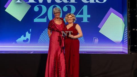 Two women, both wearing red dresses, stand in front of a blue screen. A woman with blonde hair holds a trophy in the shape of two letters, N and T. They are both smiling