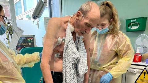 Paul Robinson is helped to stand up by nurses. He has a hospital gown hanging from his neck and has oxygen tubes in his nose.
