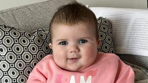 Mabli Hall sitting propped up in a grey sofa with black and grey patterned cushion. She is wearing a pink jumper.