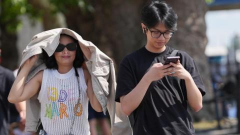 A woman holds a jacket over her head during sunny weather as a man walks next to her on his phone.
