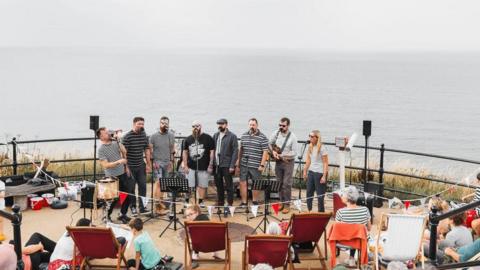Eight performers stand in a line on the promenade and sing into microphones. There is a crowd of people on the stairs and in deck chairs watching on. the sea behind them is grey and calm, and a similar colour to the sky above.