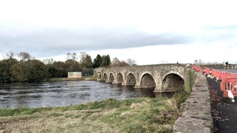 A stone bridge with large arches stretches across a river in a rural area . There are orange barriers on the bridge.