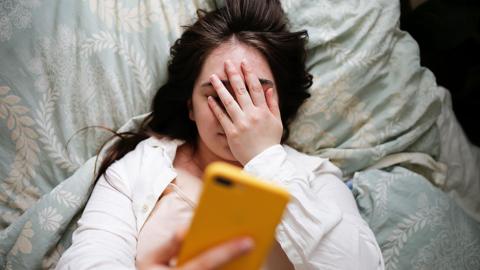 An embarrassed young woman lying down - her left hand covers her face while her right hand holds a yellow mobile phone