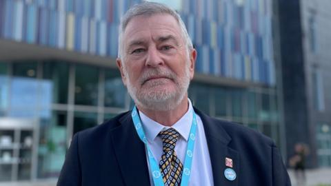 Guy Aston looks at the camera. He has short grey hair and a grey beard. He is wearing a dark suit, blue shirt and colourful tie. He has a Union Flag and Reform UK pin bad attached to his lapel.