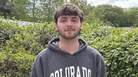 Olly has dark hair and beard and wears a dark grey hoodie with the word 'Colorado' written in white capital letters on the front. There is a hedge behind him. 