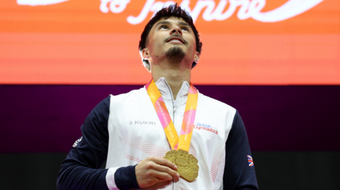 Jake Jarman is standing in the middle of the image looking up. He has short black hair and a beard and is wearing a white and navy tracksuit top. He has a gold medal around his neck. 