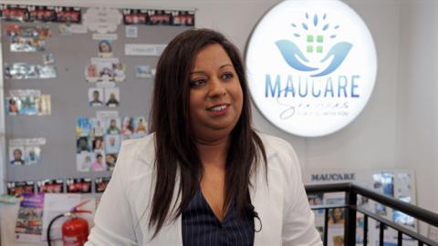 A woman in a jacket looking to the side of the image. Behind her is a display board with lots of small photographs and a large, lit up logo that reads Maucare.