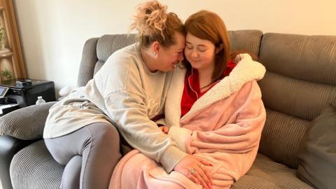 Sarah and Meadow sitting on a grey sofa. Sarah is wearing a grey sweatshirt and grey leggings. She has blonde curly hair which is tied up. Meadow is wearing a pink dressing gown. She is wearing a wig which is shoulder-length and brunette. Sarah has her head against Meadow's and smiling at her while Meadow looks down and smiles too. 