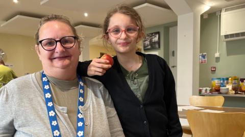 Lisa smiles at the camera as her daughter leans on her shoulder, smiling too. Lisa is sat down and wearing a grey cardigan and her daughter is wearing a black top and holding an apple. 