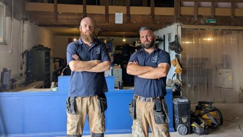 Chris Lidgerton and Ben McConnell are wearing navy blue polo necks and wearing paint splattered work trousers. They are stood with their arms crossed in front of a blue reception desk.