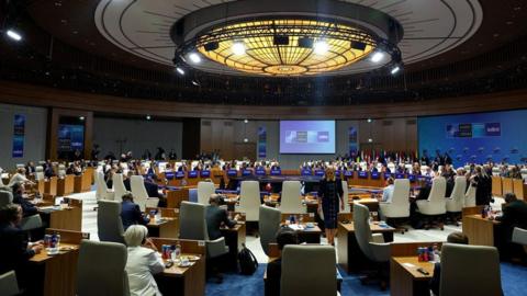 A wide view of the Nato leaders' summit in Ankara on 8 July 2026 shows delegates seated around a large circular conference table inside a modern meeting hall. Large screens display Nato branding, national flags line one side of the room and individual desks are arranged beneath a circular ceiling light.
